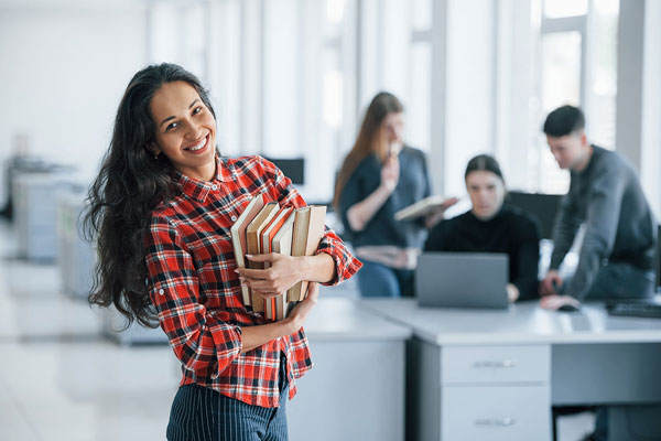 Smiling student holding a stack of books in a university study space while classmates work on laptops